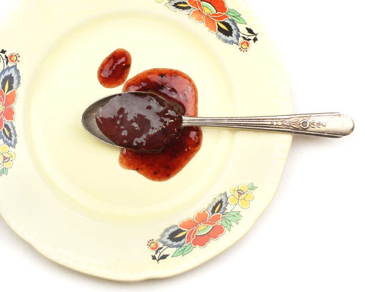 Rich Deliciousness Strawberry Limoncello Jam displayed on spoon, dark red with bursts of lemon zest, basil and black pepper